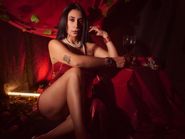 AfroditaDoir mit schwarzen Haaren, einem roten Kleid, einer Perlenkette und einem Tattoo sitzt im Schneidersitz, hält ein Glas Rotwein und ist umgeben von roten Vorhängen und Rosenblättern. Sie wirkt dunkel, sexy und verführerisch..