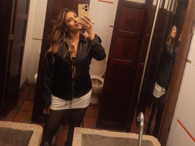 BiancaRey with long brown hair, black jacket, white mini skirt, black tights, and black boots, taking a mirror selfie, showing her ass in a bathroom.