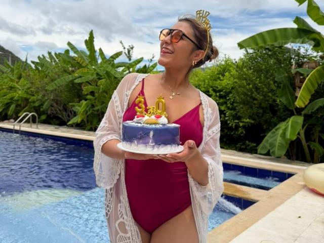 BiancaRey with big tits, wearing a red one-piece and white lace robe, holds a blue birthday cake with a "40" candle, standing by a pool, smiling.