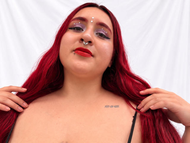 ChoeStar with red hair, nose ring, and lip ring, wearing purple eyeshadow, red lipstick, black strap top, holding hair, tattoo on right shoulder, white background.