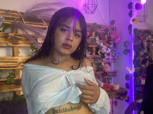 ChiaraBella with dark hair, wearing an off-shoulder white top, covering her tits with her hand, purple LED lights in the background, and a "Enchidebois" tattoo on her belly.