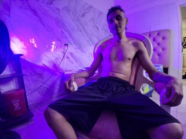 Nicolás Ferreiro, sin camisa, de complexión delgada, con pantalones cortos negros y cabello corto y oscuro, está sentado en una silla rosa, iluminado por luces moradas y azules. Su trasero es ligeramente visible..