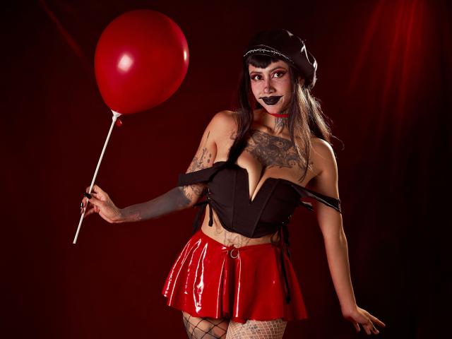 NyxVega with dark makeup, black hat, black top, red shiny skirt, fishnet stockings, holding red balloon, tattoos on arms and chest, showcasing her tits and ass.