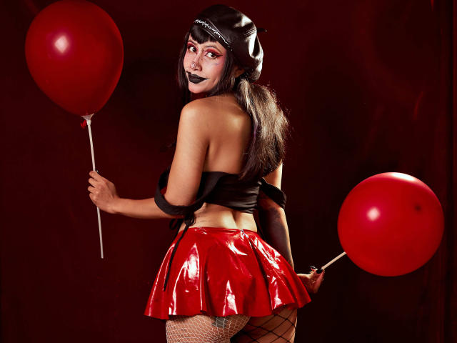 NyxVega with dark makeup, black top, shiny red skirt, fishnet stockings, holding two red balloons, looking back, wearing a black headband, in a sexy, Halloween-like outfit.