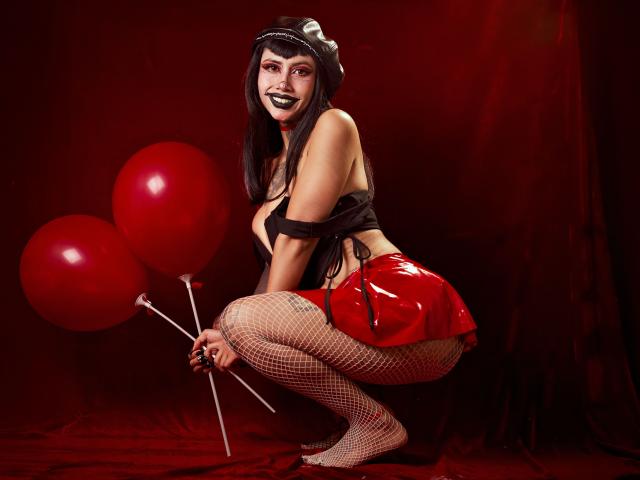 NyxVega crouching, holding red balloons, wearing black top, red shiny skirt, fishnet stockings, black lipstick, witch hat, showing tits, and a glossy red ass, against dark red backdrop.