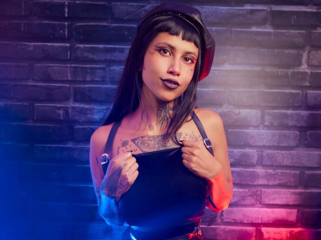 NyxVega with tattoos, black lipstick, and a leather cap, holding a black top, showing her tits, against a brick wall, with blue and red neon lights.
