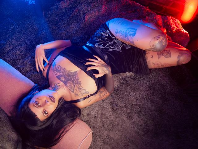NyxVega with tattoos, black dress, and thigh tattoos, lying on a carpet, legs up, arms behind head, blue and red lights, looking at camera, dark hair, red lips.