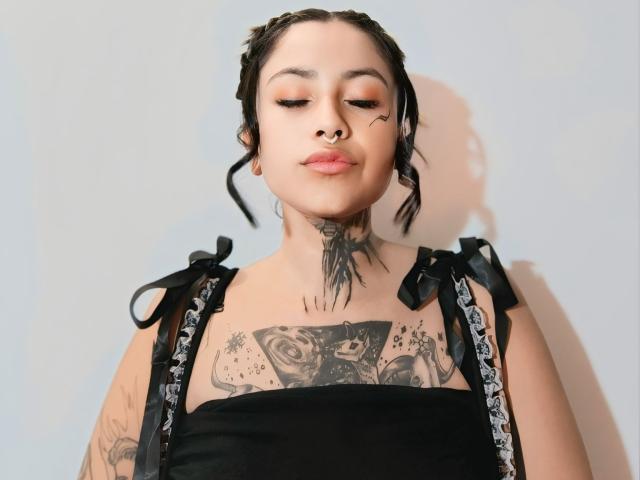 NyxVega with black braided hair, nose ring, and tattoos, wearing a black dress with ribbon straps, has pink lipstick and a confident, slightly downward gaze.