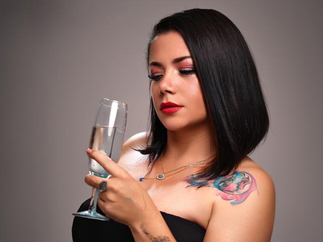 SamanthaVenus with black bob, red lipstick, holding a champagne glass, black strapless top, colorful rose tattoo on right shoulder, gray background, looking down, focused.