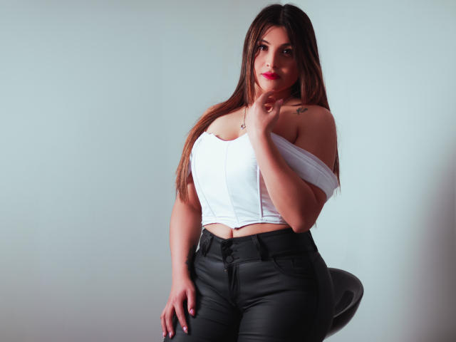 SaraWestt with long brown hair, red lipstick, white off-shoulder top, showing her tits, black high-waisted jeans, sitting on a stool, hand near her mouth, tattoo on right shoulder.