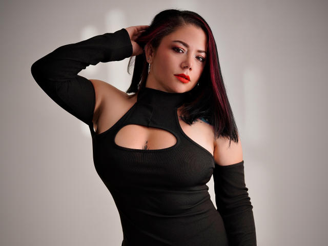 SamanthaVenus with red-black hair, wearing a black cut-out dress, showing her tits, arm raised, looking at camera, red lipstick, earrings, and a confident expression.
