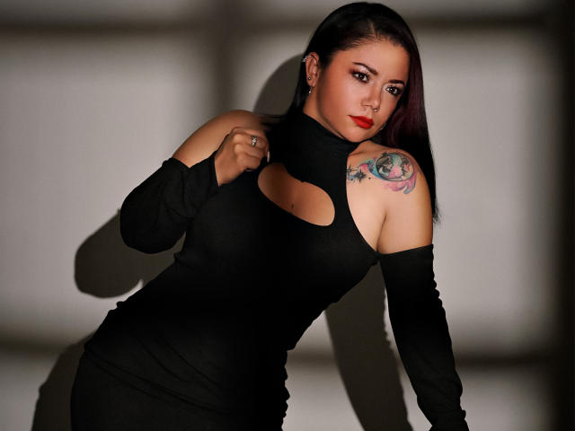 SamanthaVenus with dark hair, red lipstick, black cutout dress, and tattoo on her right shoulder, leaning forward, showing her bare shoulder and upper chest, sexy and confident.