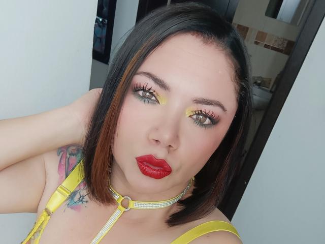 SamanthaVenus has shoulder-length black hair with brown highlights, yellow eyeshadow, red lipstick, and a yellow choker. She's wearing a yellow top, showing a tattoo on her right arm.