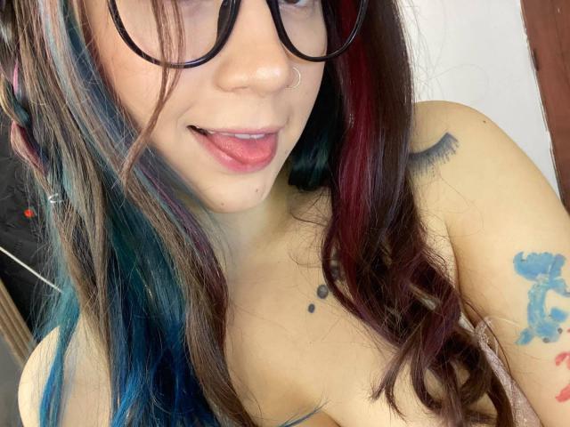 EmmaFeri with long, colorful hair, black-rimmed glasses, nose ring, tongue out, tattoos on shoulder and upper arm, and small drool on chin, topless.