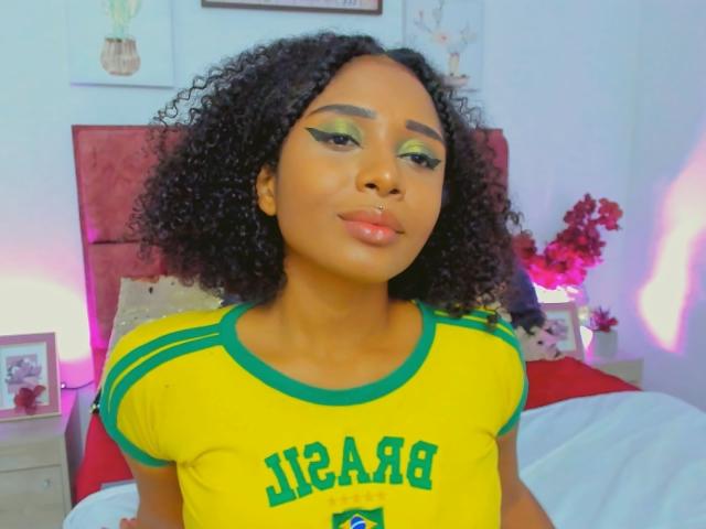 MeganWlliams with curly black hair, green and yellow "Jamaica" shirt, neon green eyeshadow, and glossy pink lips, standing in a bedroom with pink and purple lighting.