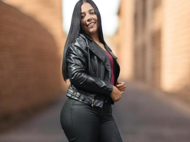 PauletteLoveX with long black hair, smiling, wearing a black leather jacket and tight black pants, showcasing her ass and curvy figure, standing in a narrow alley.