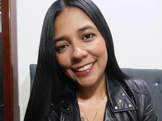 PauletteLoveX with long black hair, smiling, wearing a black leather jacket with gold studs, and a gold necklace, showing her teeth and looking at the camera.