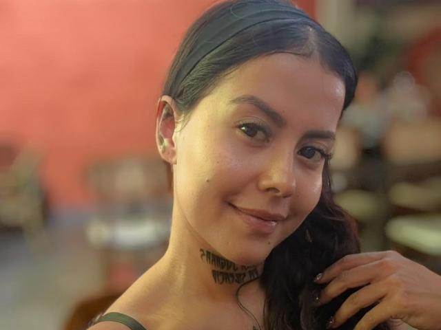 ElectraRoss has a tattoo under her chin, dark hair, and a slight smile. She's holding her hair with her right hand, showing off a cute, natural face.