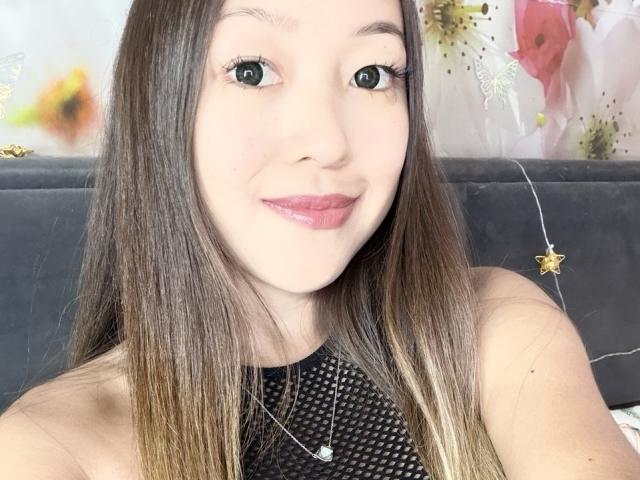 RenyLime has long brown hair, green eyes, and pink lipstick. She's wearing a black mesh top with a silver necklace. Her tits are not visible. She's smiling.