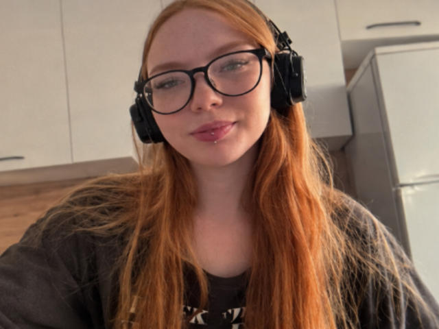 ClarineSoulia with long red hair, black glasses, and black headphones, wearing a gray shirt, has a slight smile, and pink lips. Her ass is not visible.