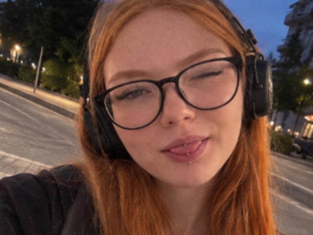 ClarineSoulia with red hair, black glasses, and black headphones, sticking out a pierced tongue, wearing a black top, in a dusk selfie.