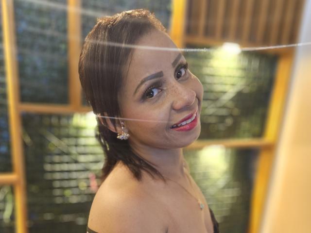 AlondraMonique has short brown hair, brown eyes, and red lipstick. She's smiling with a necklace and earrings, showing her bare shoulders and a cute, sexy face.