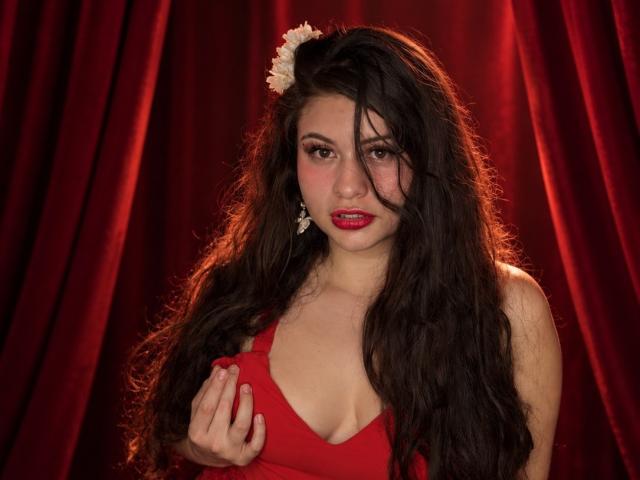 Esmaelda with long, wavy black hair, red lipstick, and a white flower in her hair, holding a red dress, showing her tits, against a red curtain.