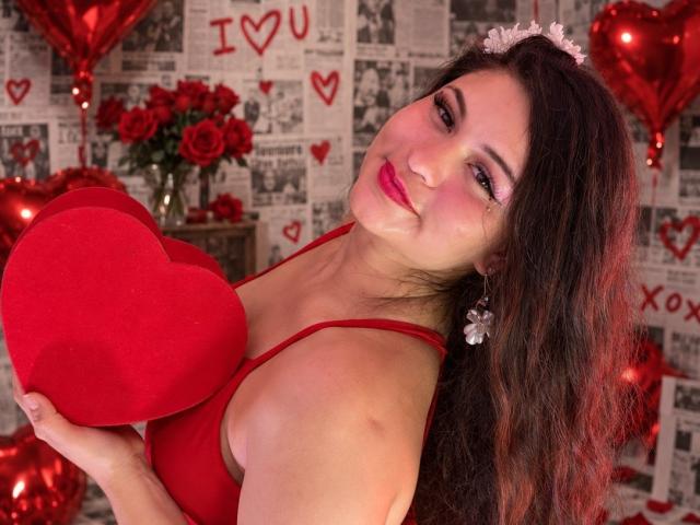 Esmaelda with red bra, holding red heart, smiling, dark wavy hair, pink eyeshadow, white flower headband, silver earrings, red lips, red balloons, roses, soft lighting.