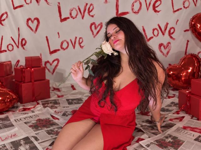 Esmaelda with long dark hair, red dress, and cleavage, holding a white rose, sitting on newspaper, surrounded by red gifts and heart balloons.