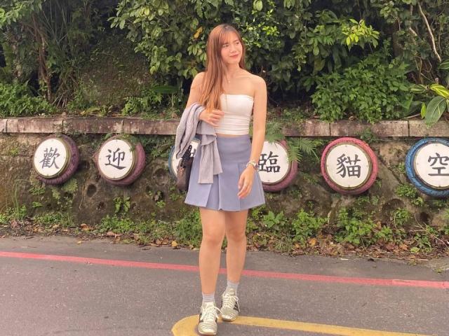 NildaReasonover, standing, has long brown hair, white tube top, blue skirt, gray jacket, sneakers, holding jacket, green leafy background, five round signs with Chinese characters.