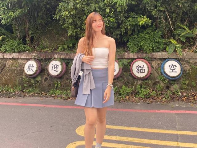 NildaReasonover with long red hair, white strapless top, blue skirt, holding gray shirt, standing on street, green leafy background, four round signs with Chinese characters.