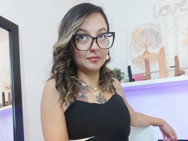 ArianaBlonde with glasses, wavy blonde and brown hair, black strapless top, showing a colorful tattoo on her chest, sitting with a relaxed smile, in a brightly lit room.