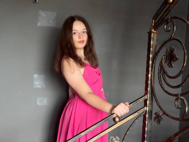 MelodiLady with wavy brown hair, pink dress, standing by a metal spiral staircase, holding the railing, smiling softly, her ass slightly visible, in a dimly lit room.
