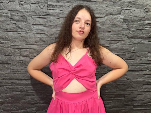 MelodiLady with wavy brown hair, pink dress, hands on hips, showing her small tits and flat ass, standing against a gray stone wall. She has a neutral expression.