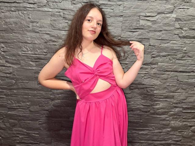 MelodiLady with long brown hair, pink dress, small tits, and a cute smile, holding her hair, standing against a gray stone wall. She's wearing a pink necklace.