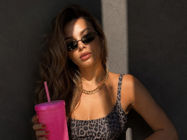 Niferttiti with long brown hair, wearing sunglasses, leopard print top, gold chain, holding pink drink with pink straw, sunlight on her face, looking relaxed.