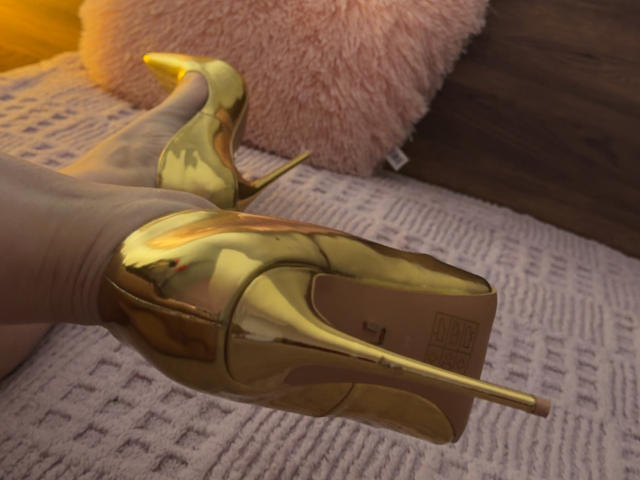 Niferttiti's shiny gold high heels are on their white-covered foot, with a pink fluffy pillow and textured gray rug in the background. Heels are pointed and reflective.