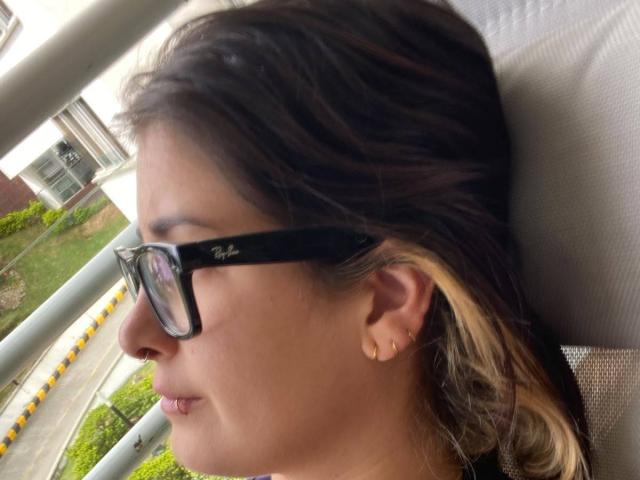 NyxVale's face, wearing black Ray-Ban glasses, nose ring, and small gold earrings, with brown and blonde ombre hair, looking outside a moving vehicle.