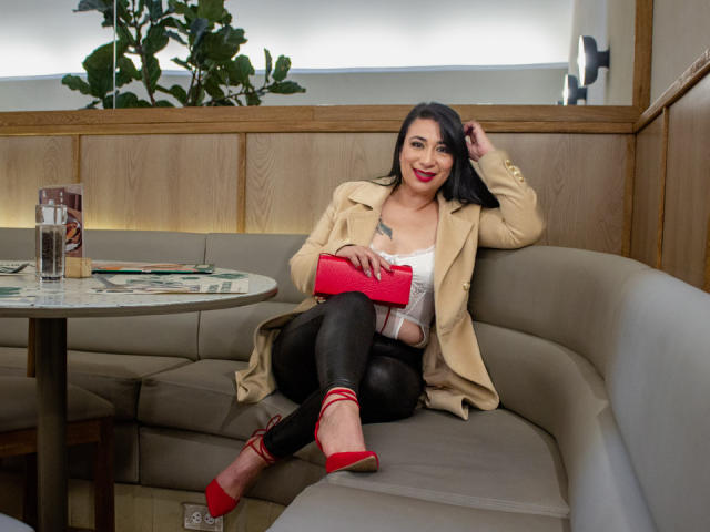Karimelee with black hair, red lipstick, beige coat, white top, black leather pants, red heels, and red clutch, sitting on gray booth, smiling, hand in hair.