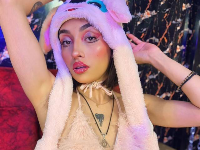 LunaixMoon with pink furry hat, glittery eyeliner, and pink lipstick, arms raised, showing tattoo on chest, wearing lace top, choker, and necklace, with pink and purple lighting.