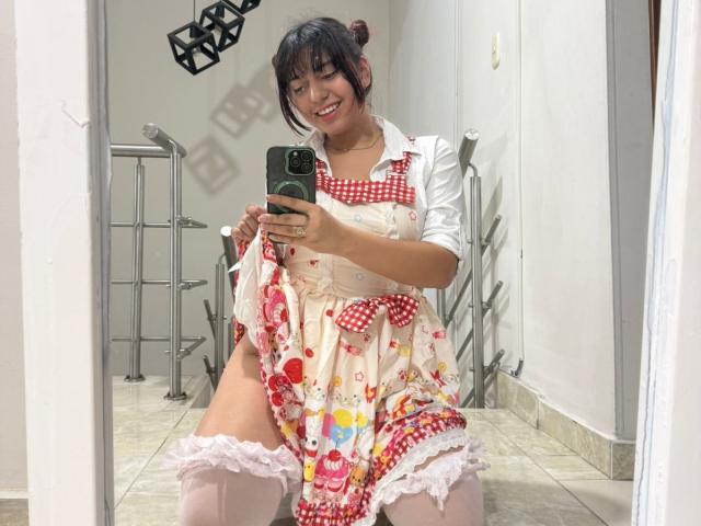 Brendaa Pierce in un carino vestito a scacchi rossi con motivi floreali, con maniche bianche a sbuffo, sorridente, che tiene un telefono, accovacciata, che mostra calze bianche e glutei scoperti, in un selfie allo specchio..