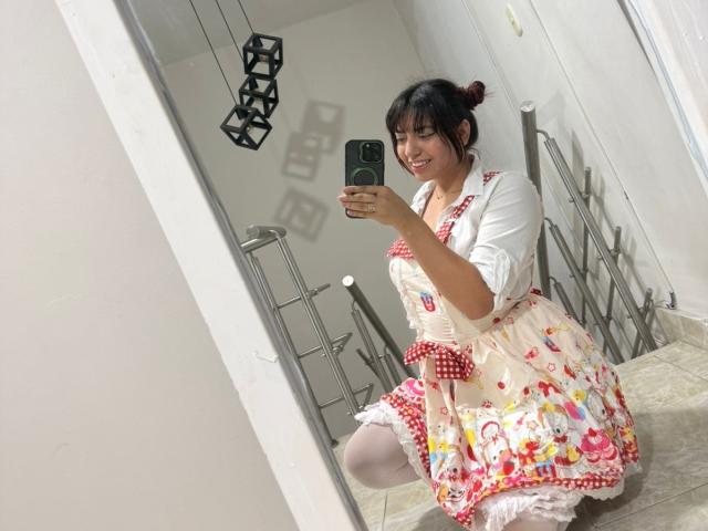 BrendaaPierce, cute Asian woman, kneeling, wearing a colorful floral apron with red polka dots, white cardigan, and white thigh-highs, taking a mirror selfie, smiling, holding a green phone.