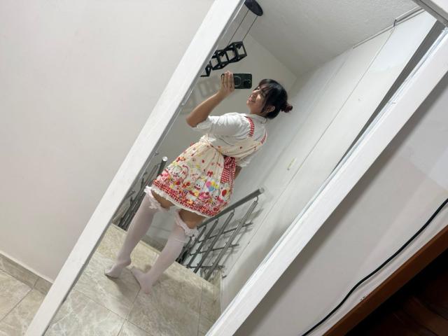 BrendaaPierce in a mirror selfie, wearing a white shirt, colorful apron, white thigh-high socks, and a bun, holding a phone, standing on a tiled floor.