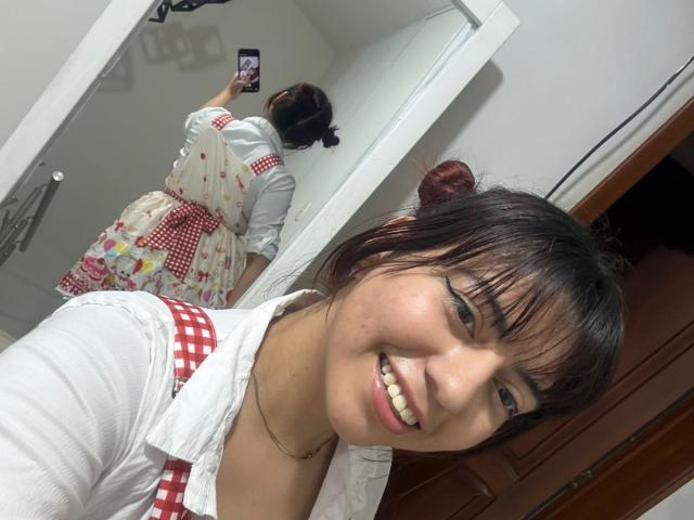 BrendaaPierce a les cheveux noirs, coiffés en deux chignons, et porte une chemise blanche avec un tablier à carreaux rouges, souriant sur une photo prise dans un miroir. Ses fesses sont couvertes par un tablier fleuri..