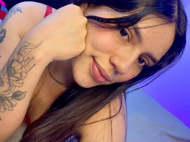 SophiaMarx with long brown hair, smiling, resting chin on hand, showing tattoo on her arm, pink nipples visible, wearing red top, blue-purple light, bed in background.