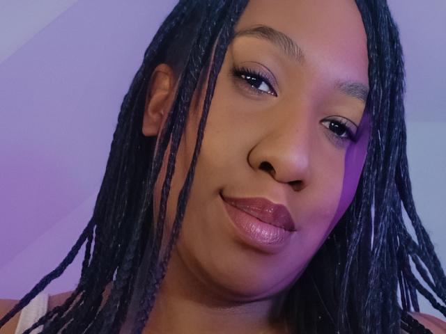 Luxnyx a les cheveux noirs tressés, une peau brune et lisse, et des lèvres pulpeuses avec un rouge à lèvres rose brillant. Elle regarde légèrement sur le côté avec un sourire discret..
