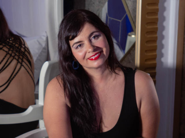 ScarletRebel, a curvy woman with long dark brown hair, red lipstick, and blue earrings, wearing a black tank top, smiling at the camera, showing her medium-sized boobs.