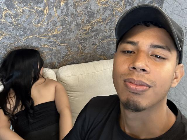 SofiaAndAndres has brown skin, a black cap, and a black shirt. SofiaAndAndres's butt is covered in a black strapless top, lying on a couch.
