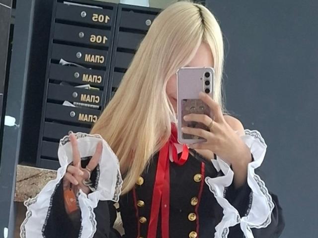JanettaKnop with blonde hair, wearing a black and white cosplay outfit with red ribbon, holding a phone in front of her face, showing a finger gesture.