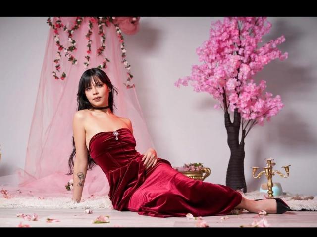MadameDesadex with black hair, red velvet strapless dress, tattoo on left arm, sitting on pink cloth, pink flowers, pink tree, and gold lamp in soft pink room.